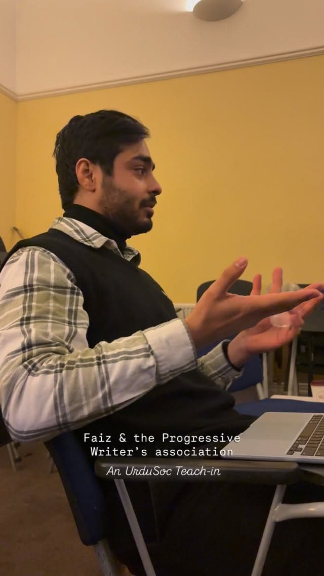 A speaker mid-sentence in a yellow-walled seminar room, hands raised, a laptop open on the desk beside him. The frame is captioned 'Faiz & the Progressive Writer's association - An UrduSoc Teach-in'.