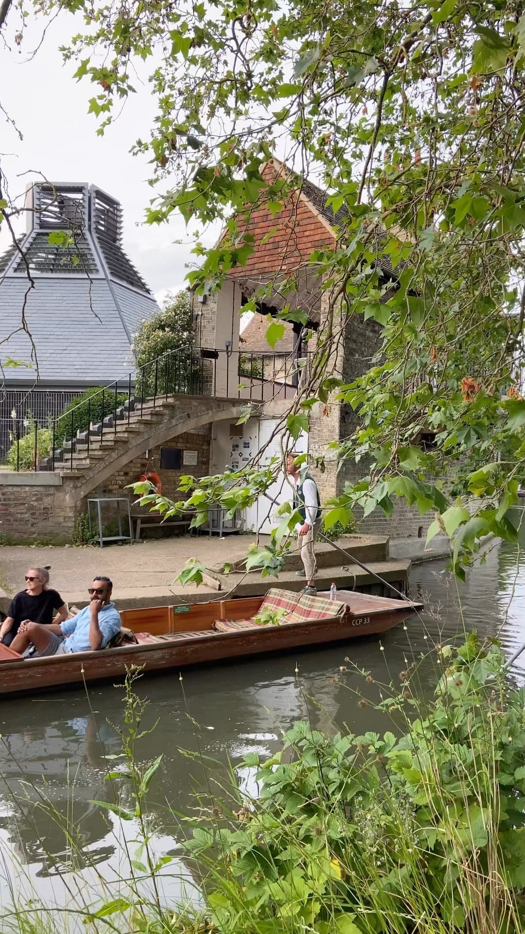 A punt drawing in beside a boathouse on the Cam, two passengers seated in the bow and the chauffeur standing at the stern with the pole, the view framed by overhanging plane leaves.